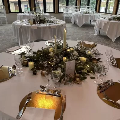 Salle de réception soigneusement aménagée avec des tables rondes, des nappes blanches, des compositions florales, des bougies et des couverts élégants