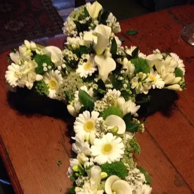 Arrangement floral en forme de croix composé de marguerites blanches, de lys blancs et de verdure, disposé sur une table en bois