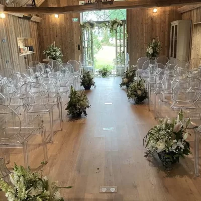 Espace intérieur en bois avec des poutres apparentes, prêt pour une cérémonie. Chaises transparentes alignées de chaque côté d'un couloir central, orné de belles compositions florales. Une porte vitrée ouverte offre une vue sur l'extérieur